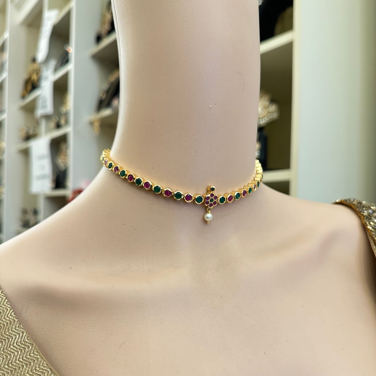 A gold-plated choker necklace with green and red gemstone accents and a central pearl-like drop,  jewelry store.