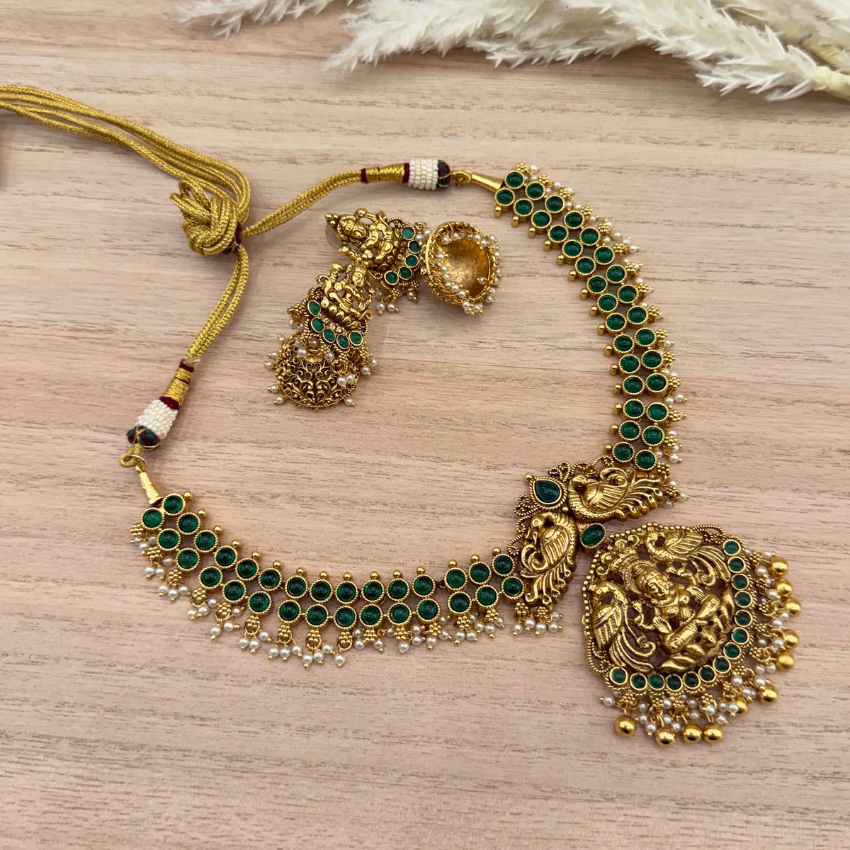 Traditional gold-tone temple jewellery necklace set with emerald green stones, intricate goddess design, matching earrings, and pearl detailing. Available in emerald green and white stone options.
