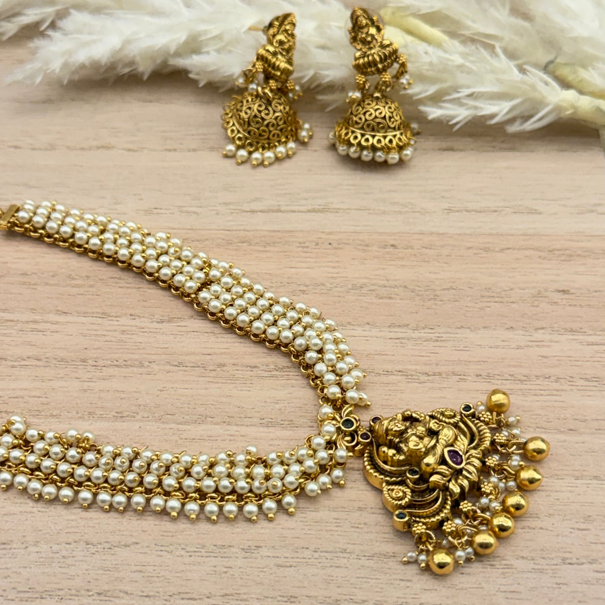 Traditional South Indian antique gold-tone temple jewellery necklace set featuring a detailed deity pendant, multiple rows of white pearls, gold bead drops, and matching jhumka earrings.