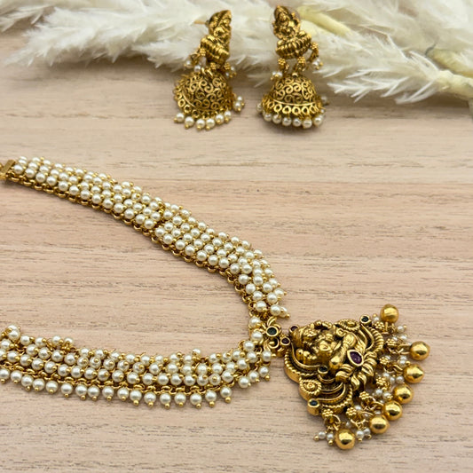 Traditional South Indian antique gold-tone temple jewellery necklace set featuring a detailed deity pendant, multiple rows of white pearls, gold bead drops, and matching jhumka earrings.